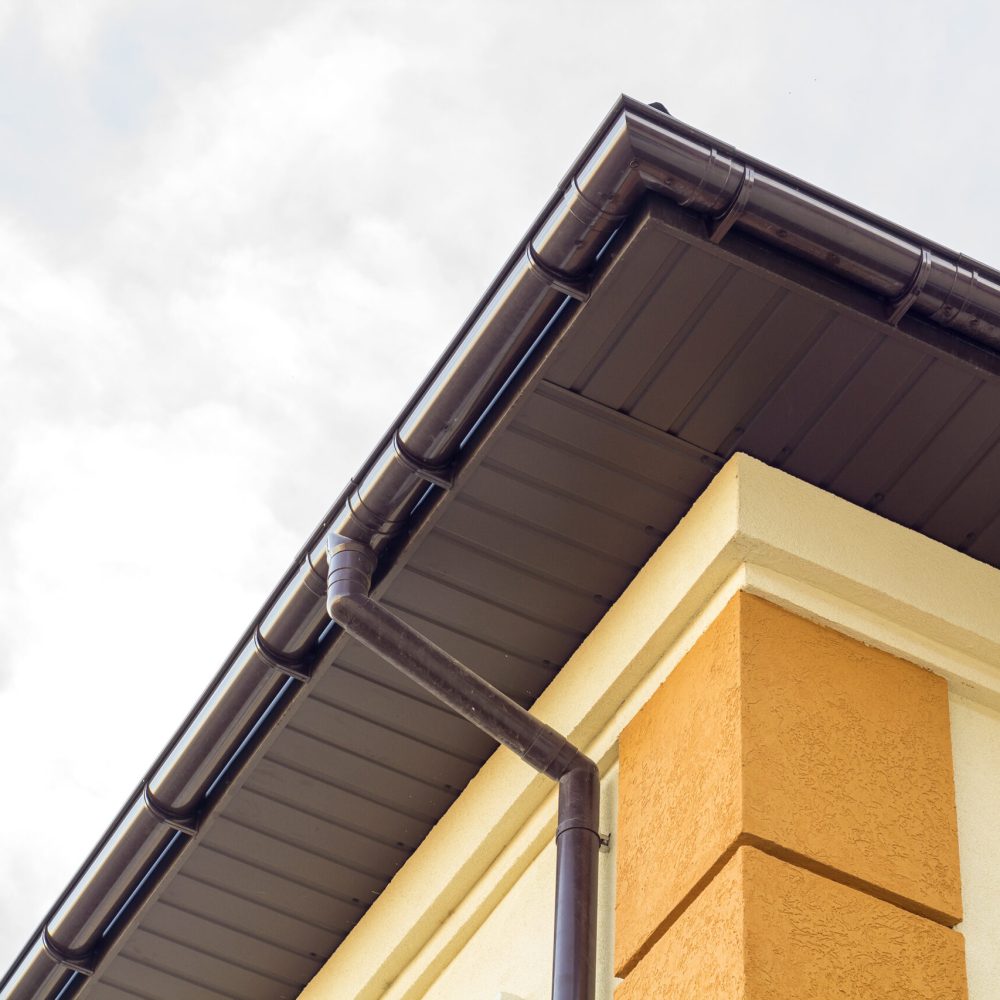 Close-up detail of cottage house corner with brown metal planks siding and roof with steel gutter rain system. Roofing, construction, drainage pipes installation and connection concept.